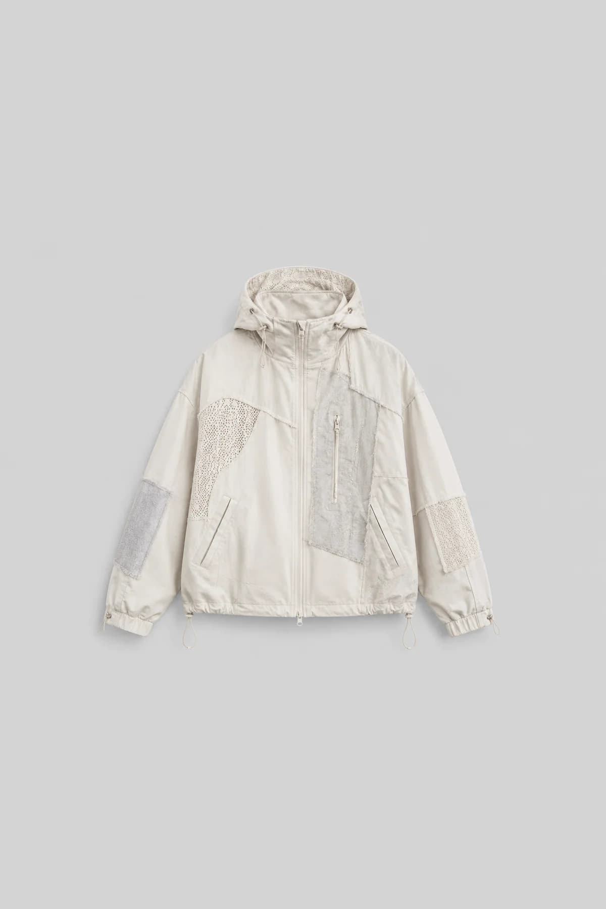 Stone-grey hooded zip anorak laid flat, mesh and fabric patches pieced across the chest and sleeves