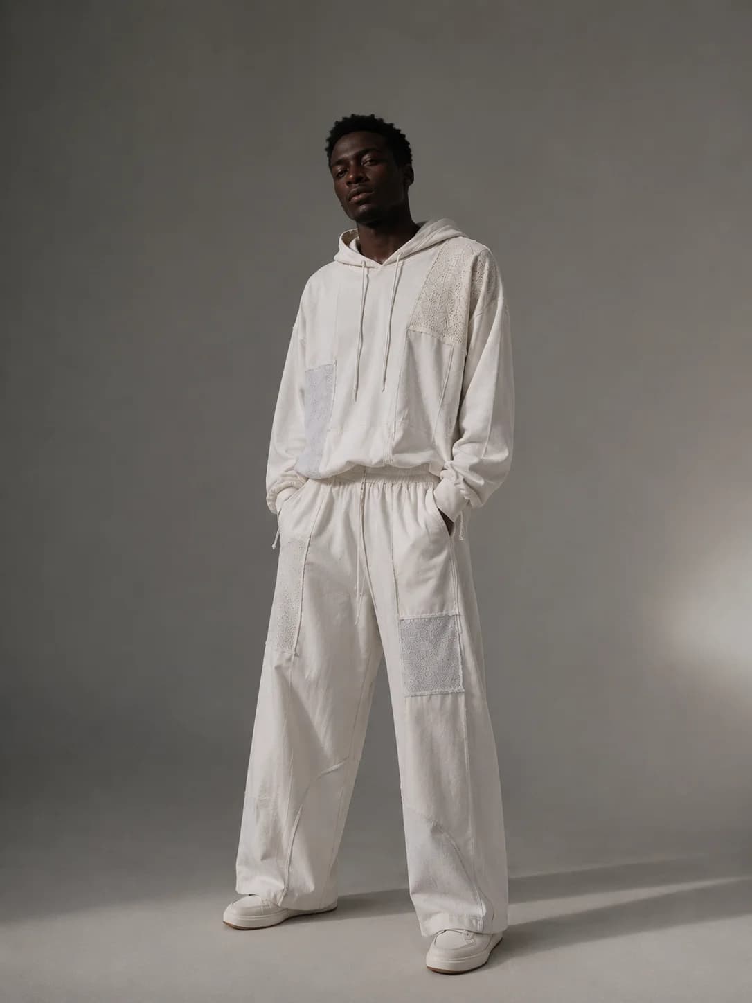 A man in the ivory patchwork hoodie and matching wide trousers against a grey studio backdrop
