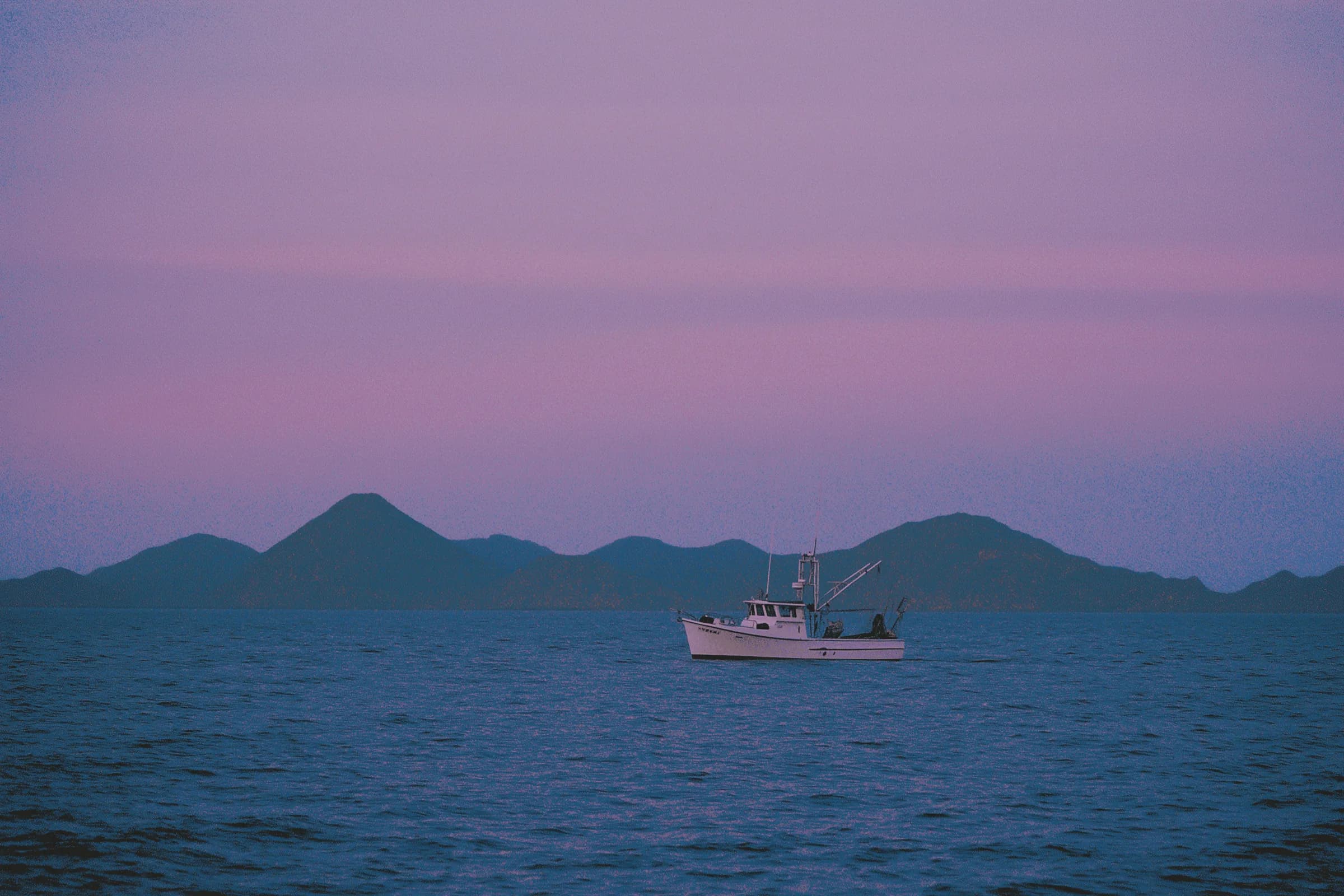A fishing boat on a calm blue ocean, hills and a pink-purple sunset sky in the background.